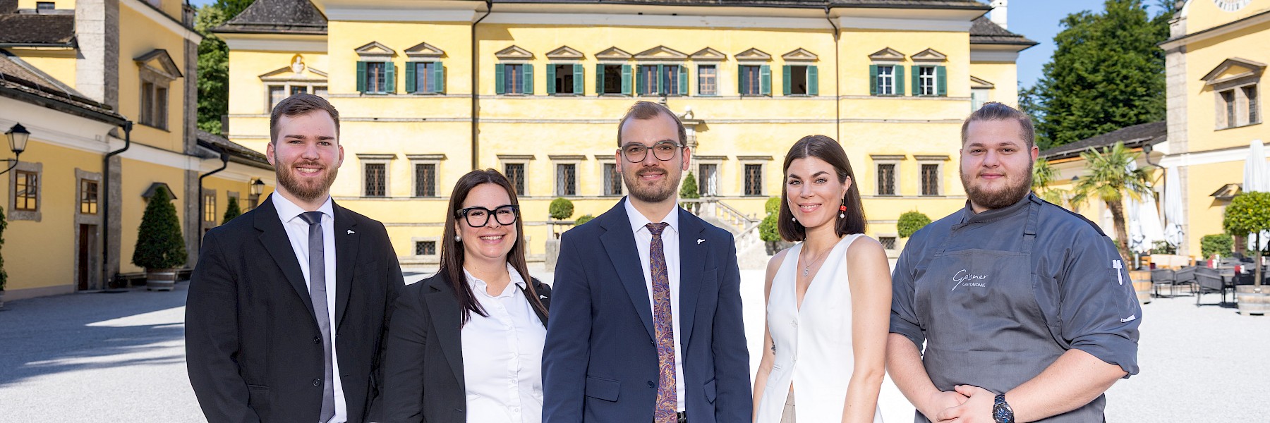 Vor der historischen Kulisse von Schloss Hellbrunn präsentiert sich ein Teil des Teams des Gasthauses in eleganter und professioneller Erscheinung. Fünf Teammitglieder stehen nebeneinander – drei in stilvollen Anzügen, eine in sommerlich-leichter Businesskleidung und einer in moderner Küchenuniform. Alle strahlen Herzlichkeit, Kompetenz und Teamgeist aus. Das gelbe Schlossgebäude im Hintergrund und der blaue Himmel verleihen dem Bild eine warme, einladende Atmosphäre. Dieses Foto spiegelt die perfekte Verbindung von Tradition, Gastfreundschaft und moderner Gastronomiekultur wider – das Herzstück des Gasthauses zu Schloss Hellbrunn.