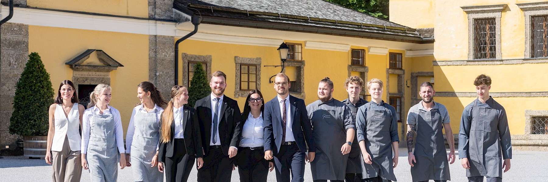 Gasthaus Hellbrunn, Foto Streb: The image shows the friendly and professional team of the Gasthaus at Schloss Hellbrunn, consisting of twelve staff members. They are standing in an elegantly set dining room with tall windows and green curtains. Some team members wear gray aprons with the Gasthaus logo, while others are dressed in classic suits with ties. Everyone is smiling warmly at the camera, creating a welcoming and positive impression. The scene conveys a strong sense of teamwork, hospitality, and dedication to excellent service—an ideal team ready to offer guests a memorable culinary experience in a refined atmosphere.