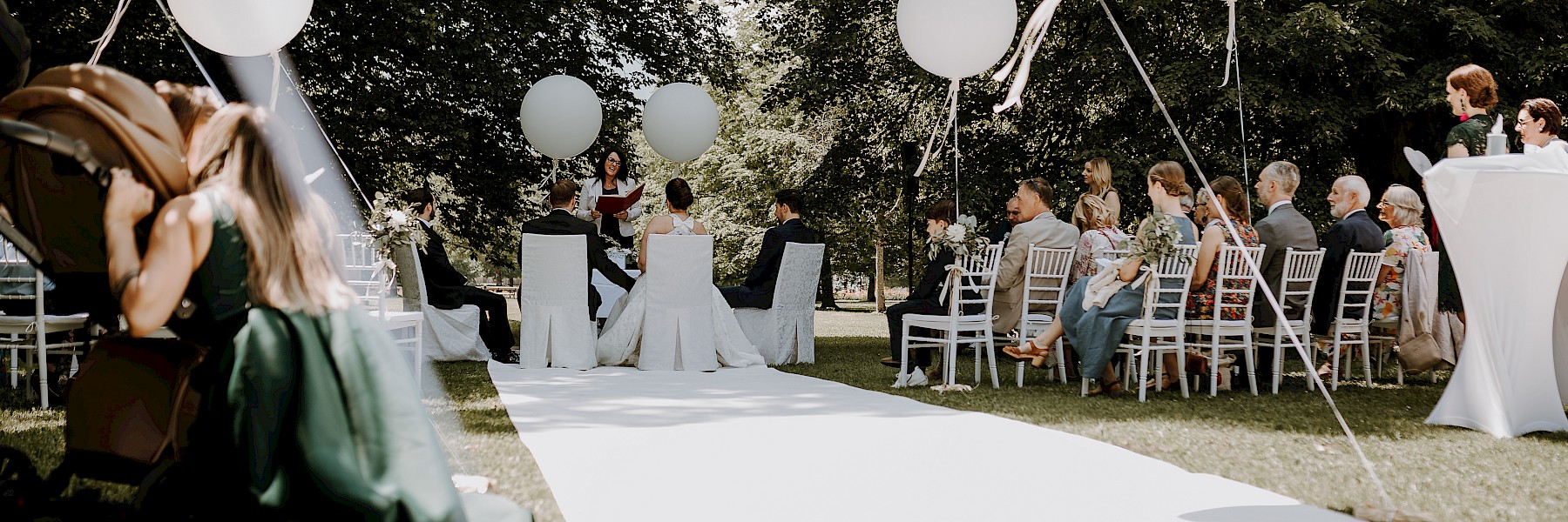 Heiraten in Hellbrunn (c) Gassner Gastronomie, Fotografie Farbenspiel: The image captures a newlywed couple standing hand in hand in front of a serene body of water. The bride is wearing an elegant, sleeveless white gown with a veil and holding a bouquet of white roses and greenery. The groom is dressed in a classic black tuxedo with a bow tie and a boutonniere. They are smiling at each other warmly. In the background, beautifully manicured gardens, a charming pavilion, and a stately yellow château enhance the romantic, fairytale-like atmosphere—perfectly reflecting the joy and elegance of their special day.