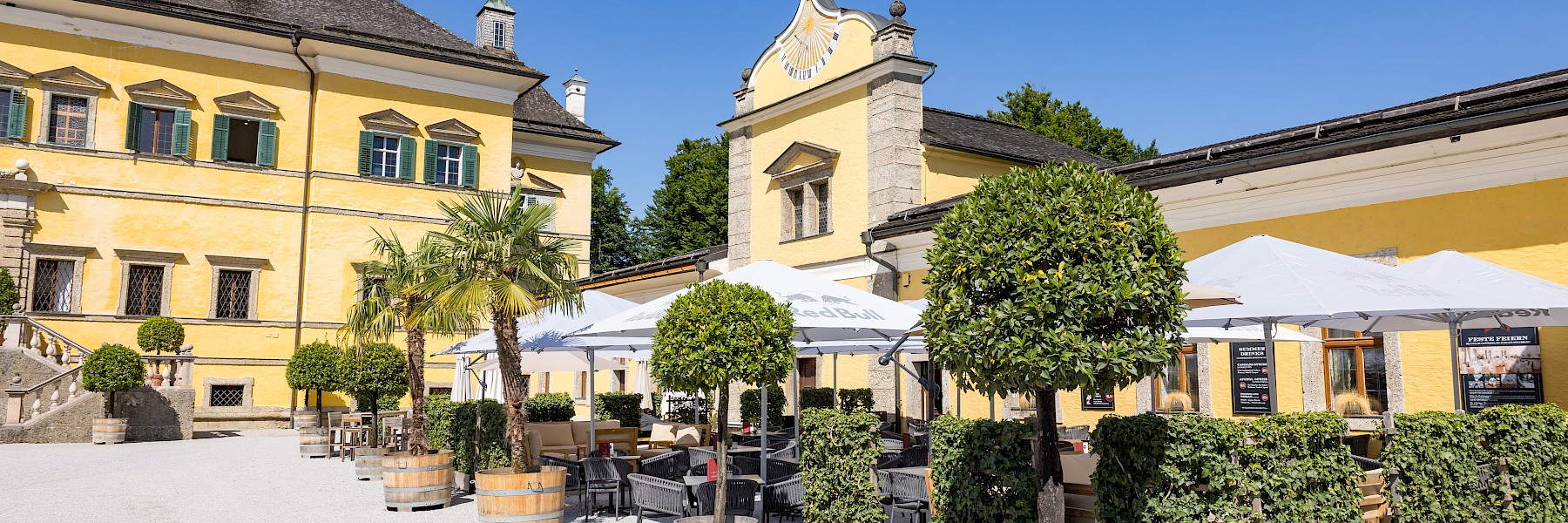 Hellbrunn Schloss (c) Schlossverwaltung Hellbrunn Foto Auer:  The image shows the impressive façade of Hellbrunn Palace in Salzburg, a Baroque pleasure palace painted in a warm yellow with green shutters. In front of the grand entrance lies a neatly maintained courtyard with a rectangular reflecting pool, framed by stone statues and symmetrically arranged plants. A group of visitors gathers at the entrance, adding life to the serene setting. Surrounding the palace are manicured hedges, lush lawns, and ornamental garden features. In the background, forested hills rise under a clear blue sky, creating a picturesque and elegant scene rich in historical charm.